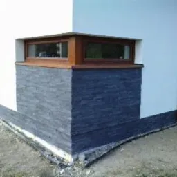 Wykończony narożnik budynku z oknem, z dolną częścią elewacji pokrytą szarym kamieniem dekoracyjnym i białą, gładką ścianą powyżej. Widoczne obrzeża i fragment trawnika.