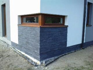 Wykończony narożnik budynku z oknem, z dolną częścią elewacji pokrytą szarym kamieniem dekoracyjnym i białą, gładką ścianą powyżej. Widoczne obrzeża i fragment trawnika.