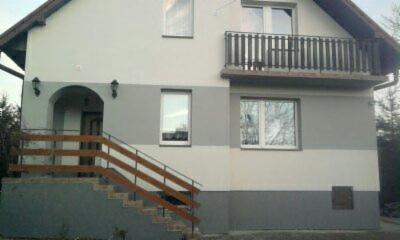 Dwukolorowa elewacja domu jednorodzinnego z drewnianą balustradą schodów wejściowych i balkonem z ciemnymi barierkami.