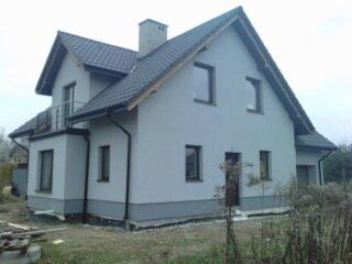 Dwukondygnacyjny dom jednorodzinny z szarą elewacją i ciemnym dachem, widok z boku, z widocznym fragmentem balkonu i komina.