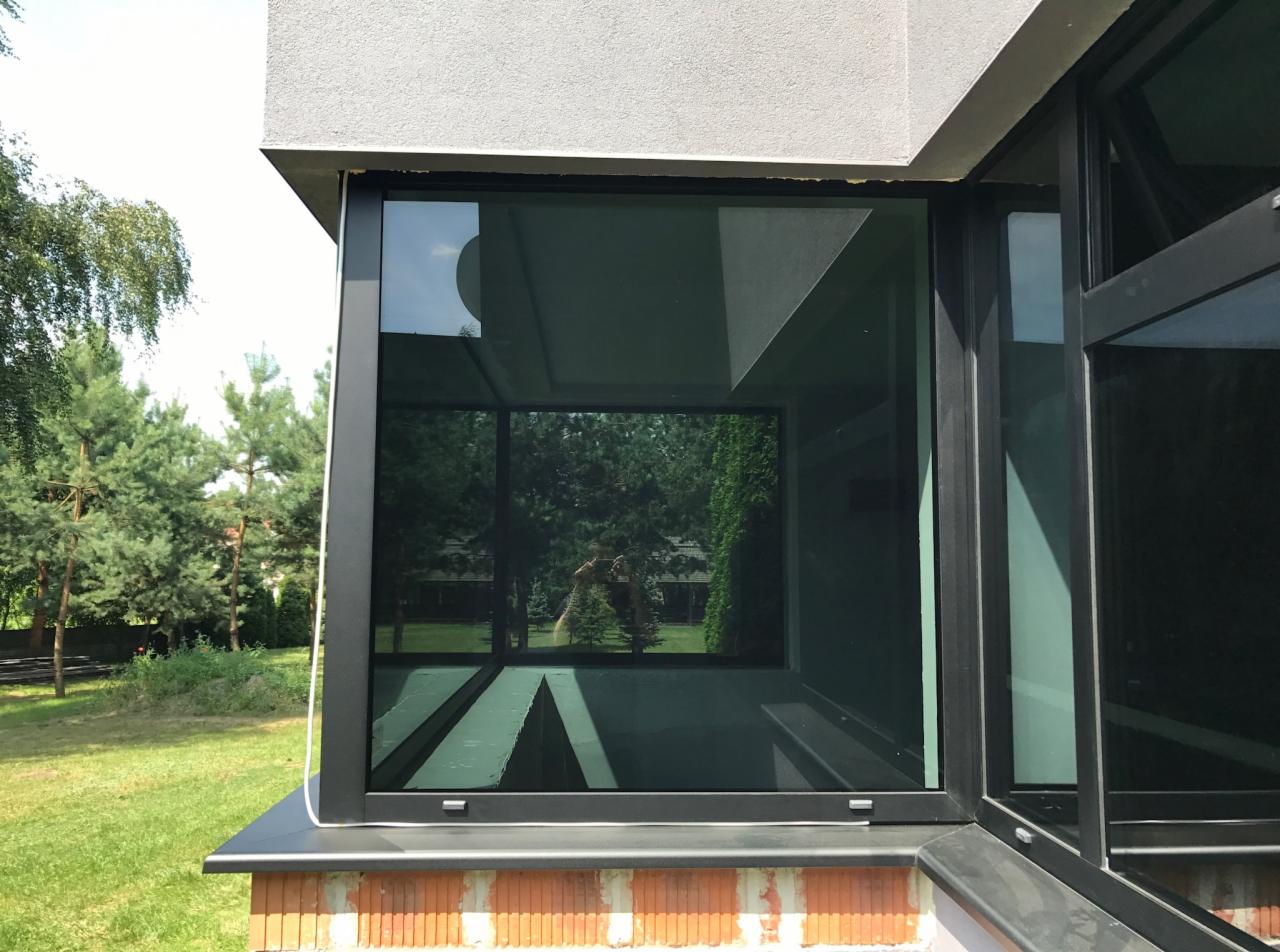 Narożne okno aluminiowe z czarną ramą, zamontowane na murze z cegły, z widocznym odbiciem ogrodu i osoby robiącej zdjęcie.