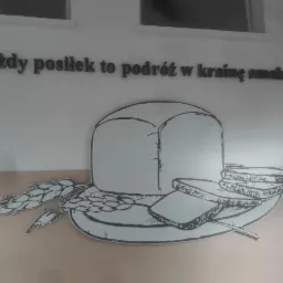 Grafika ścienna przedstawiająca bochenek chleba, kromki i kłosy zboża na talerzu, nad nią napis 'Każdy posiłek to podróż w krainę smaku...'