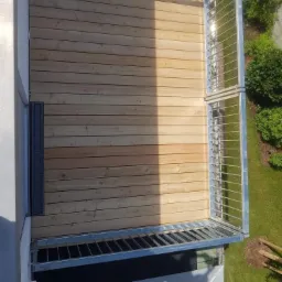 Nowoczesny balkon z jasną, drewnianą podłogą ułożoną z desek, metalowa balustrada z cienkich prętów, widok z góry na fragment zielonego trawnika i krzewów w tle.