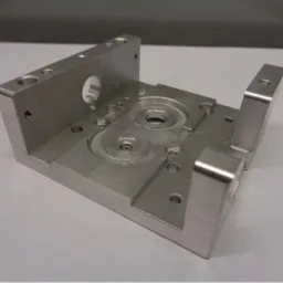 Precyzyjnie obrobiony, surowy, aluminiowy element konstrukcyjny z licznymi otworami i wgłębieniami, ukazujący ślady frezowania i gwintowania, leżący na jasnym tle.