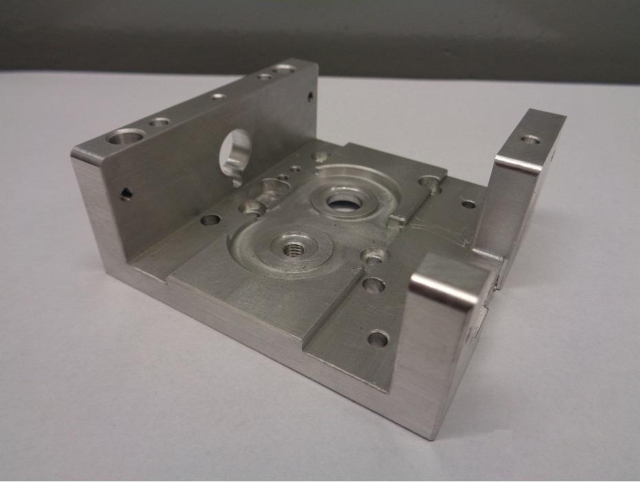 Precyzyjnie obrobiony, surowy, aluminiowy element konstrukcyjny z licznymi otworami i wgłębieniami, ukazujący ślady frezowania i gwintowania, leżący na jasnym tle.