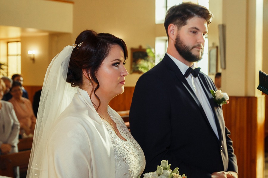 Para młoda podczas ceremonii ślubnej w kościele, panna młoda w koronkowej sukni i welonie, pan młody w granatowym garniturze i muszce.