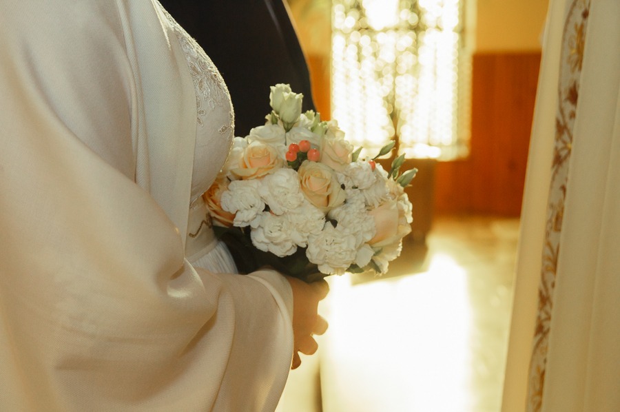 Panna młoda w kremowej sukni trzyma bukiet z białych goździków i brzoskwiniowych róż na tle okna z rozproszonym światłem.