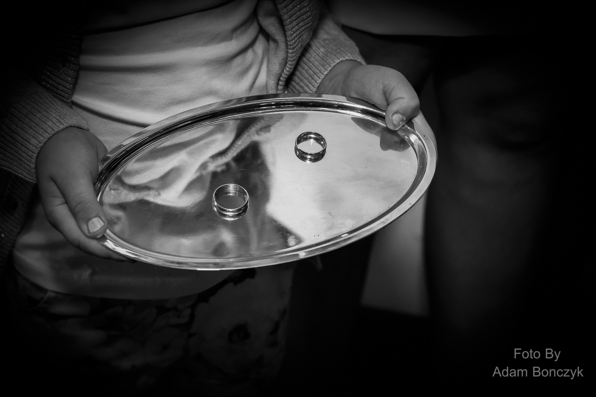 Dziecięce dłonie trzymają srebrną tacę z dwoma obrączkami ślubnymi, czarno-biała fotografia.