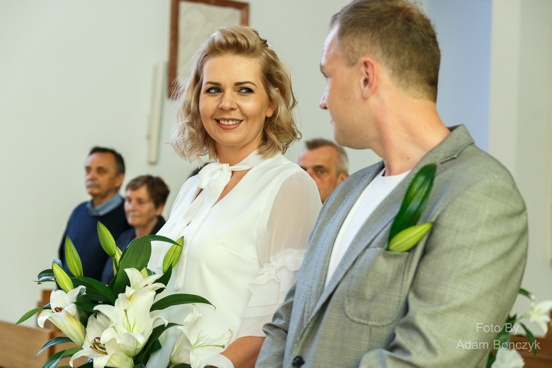 Uśmiechnięta blond kobieta w białej bluzce z bukietem lilii spogląda na mężczyznę w szarej marynarce z zielonym pąkiem lilii w butonierce, w tle widoczni goście.