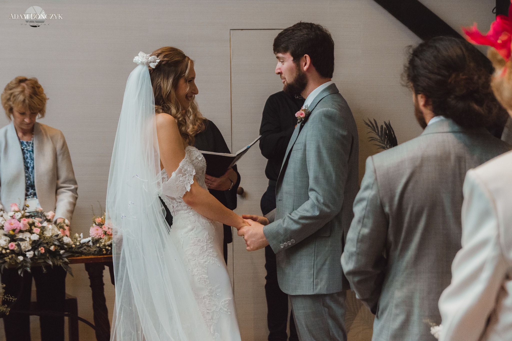 Para młoda trzymająca się za ręce podczas ceremonii ślubnej, panna młoda w białej sukni z koronką i welonem, pan młody w szarym garniturze z butonierką, w tle kwiaty i goście.