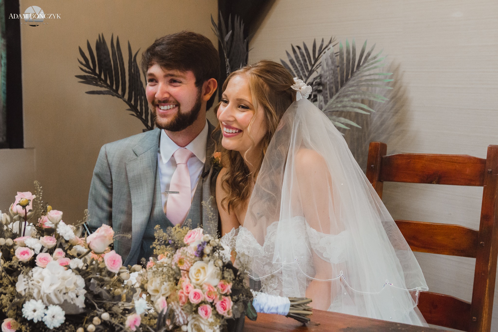 Uśmiechnięta para młoda siedząca za stołem udekorowanym kwiatami podczas ceremonii ślubnej, widoczne kwiaty we włosach panny młodej i delikatny welon.