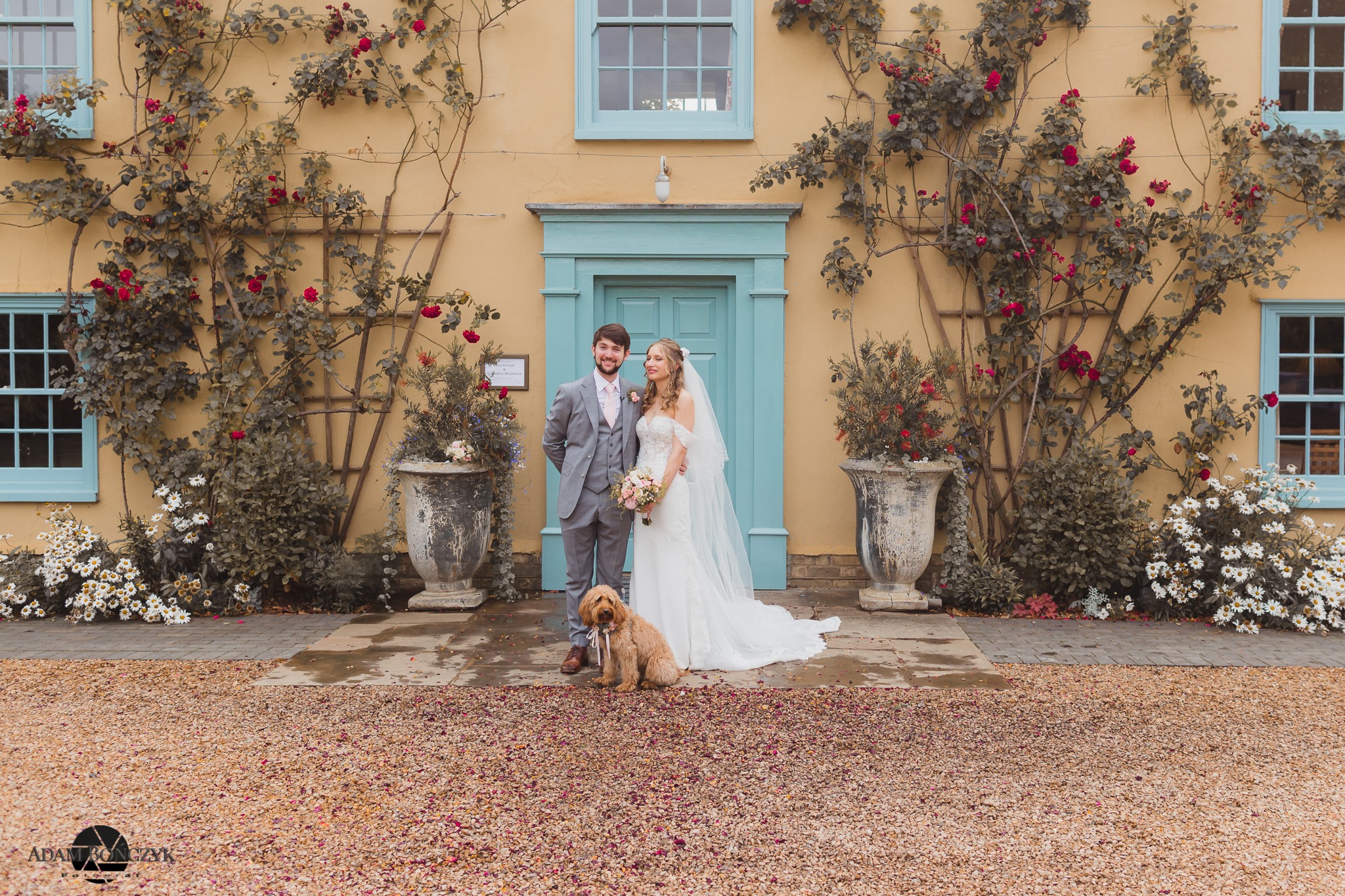 Para młoda w eleganckich strojach ślubnych stoi przed żółtym budynkiem z turkusowymi oknami i drzwiami, otoczonym pnącymi różami, a u ich stóp siedzi pies z kokardą.