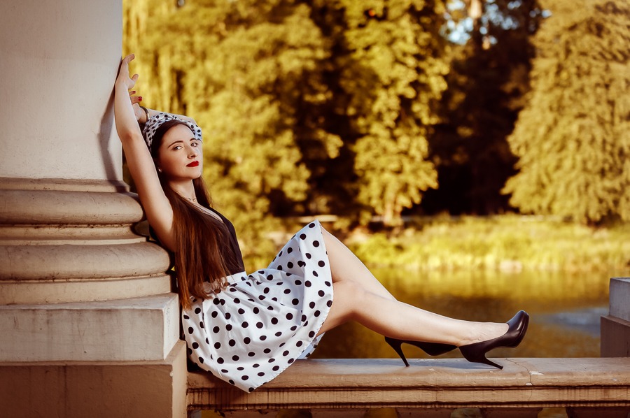 Kobieta w sukience w grochy i czarnych szpilkach pozuje oparta o kolumnę na tle parku w Kaliszu, w stylu retro.