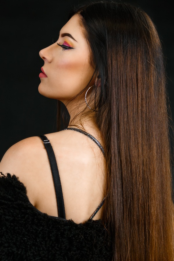 Portret kobiety z długimi, prostymi brązowymi włosami, widok z profilu, z wyrazistym makijażem oczu w odcieniach różu, pomarańczy i żółci, srebrne kolczyki koła, czarny sweter i ramiączka biustonosza