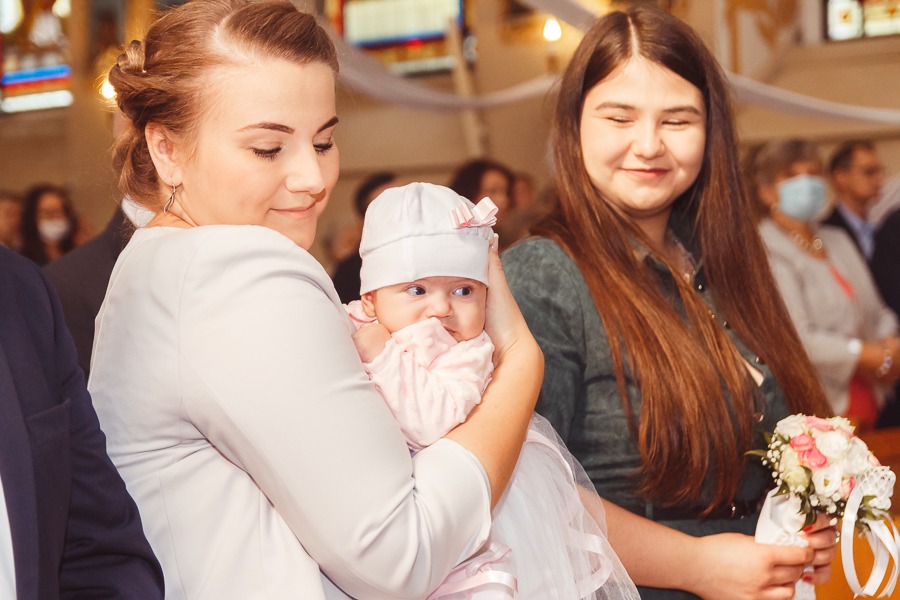 Matka trzymająca na rękach ubraną na różowo dziewczynkę w białym czepku z kokardką, w tle kościół i kobieta z bukietem.