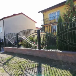 Czarne, metalowe ogrodzenie z falistym wzorem na niskim, ceglanym murku, przed domami w słoneczny dzień.