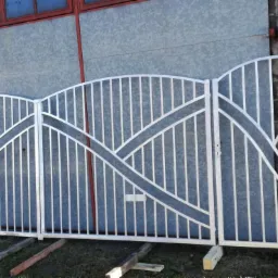 Trzyczęściowa, srebrna brama z metalowymi prętami i falistymi elementami dekoracyjnymi, oparta na drewnianych klockach, na tle szarej ściany budynku i muru z szarych cegieł.