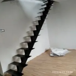 Schody wspornikowe z czarnego metalu, stopnie mocowane do ściany, widoczne ślady prac wykończeniowych na podłodze, surowe wnętrze.