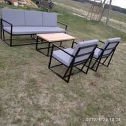 Metalowy zestaw mebli ogrodowych: sofa trzyosobowa, dwa fotele i stolik kawowy z drewnianym blatem, na trawniku, widok z boku, konstrukcja spawana z profili zamkniętych.