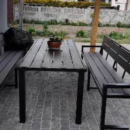 Zestaw mebli ogrodowych: stół z blatem imitującym drewno i metalowymi nogami, dwie ławki z oparciami i podłokietnikami, donica z kwiatami na stole, całość na brukowanej powierzchni, w tle...