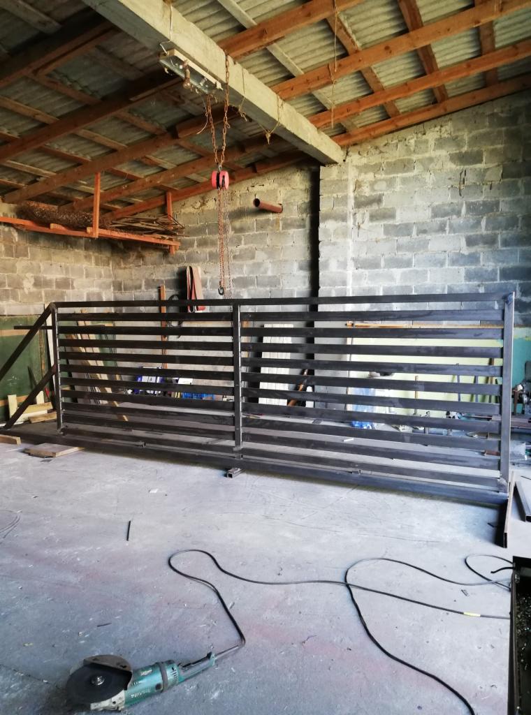 Surowa, metalowa brama wjazdowa z poziomymi szczeblami, w trakcie produkcji w warsztacie, widoczna szlifierka kątowa na podłodze i suwnica łańcuchowa pod drewnianym dachem.