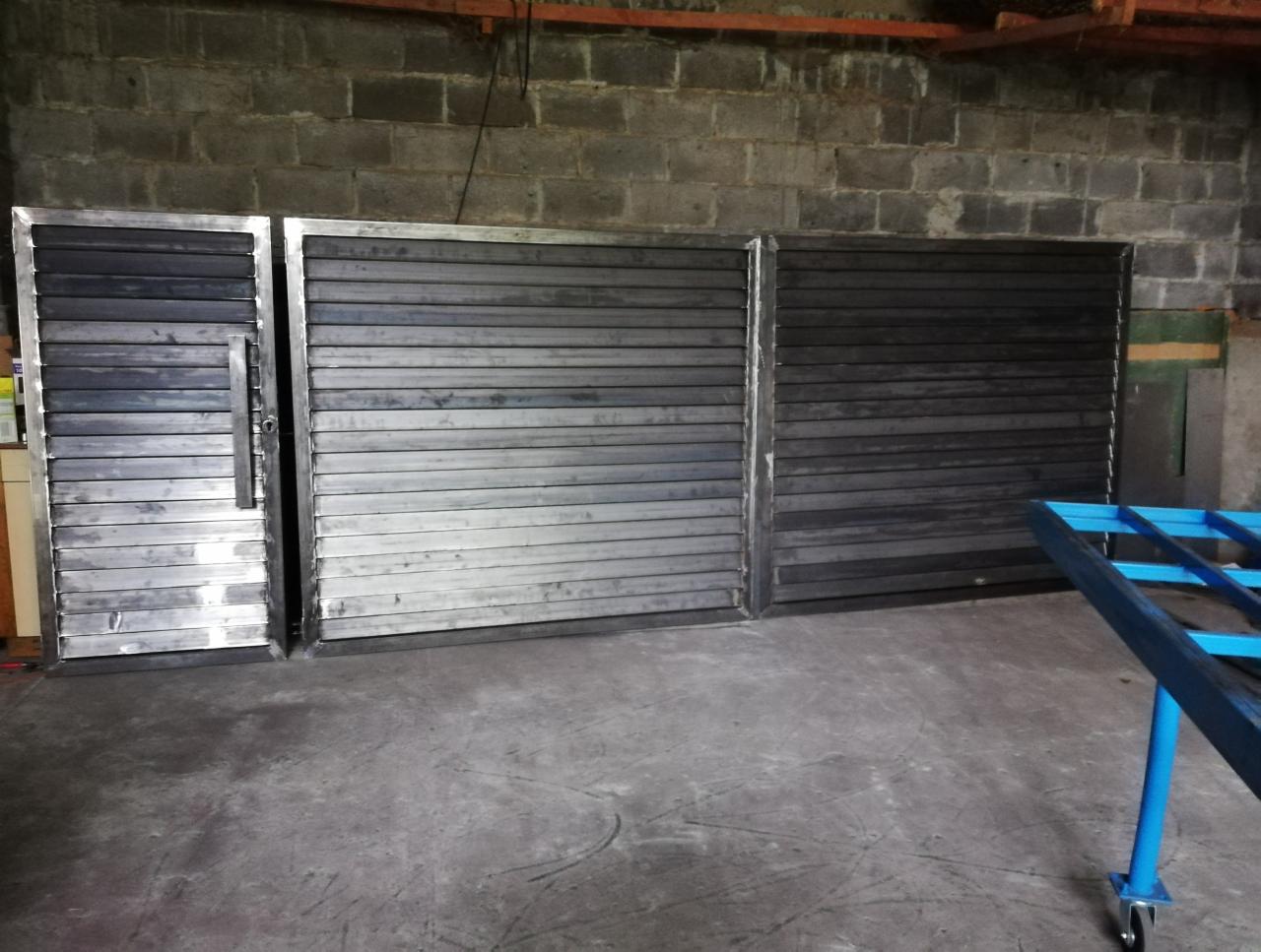 Surowe, metalowe bramy wjazdowe z poziomymi szczeblami, przygotowane do montażu, w warsztacie z ceglaną ścianą w tle.
