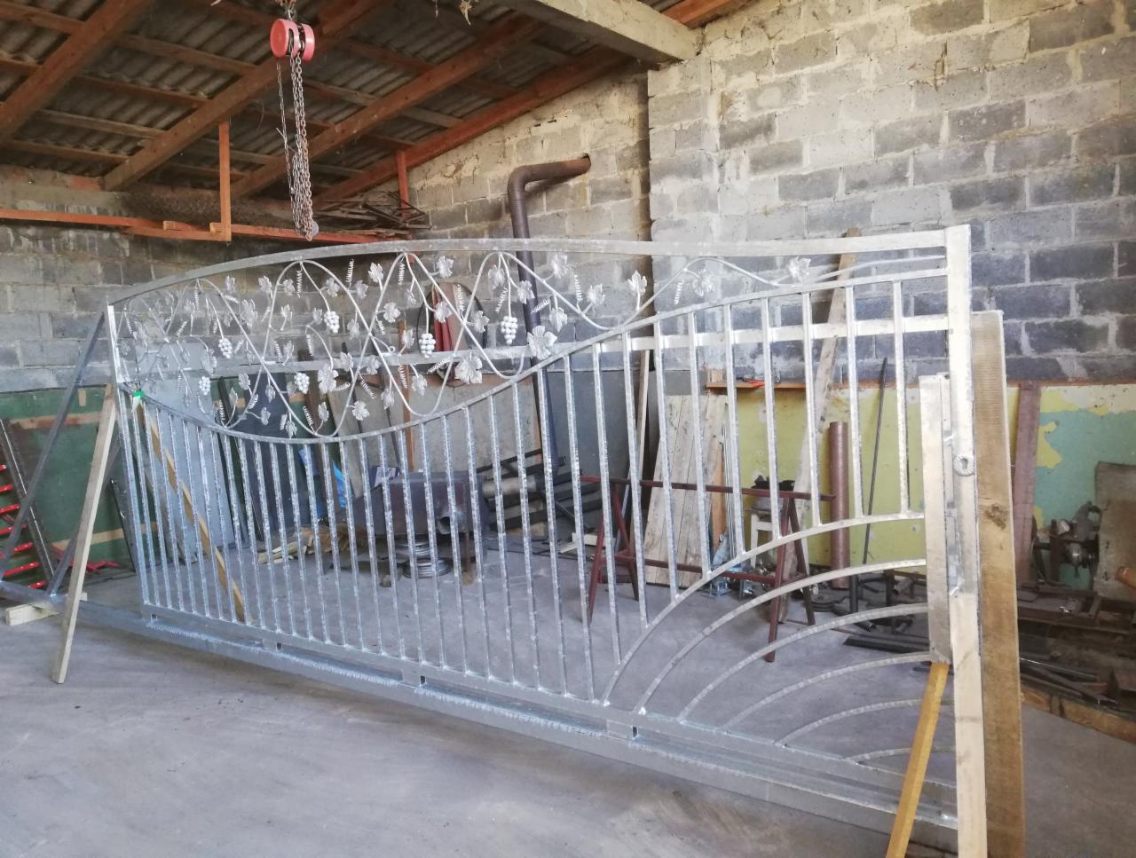 Surowa, metalowa brama wjazdowa z motywem winogron, podparta drewnianymi belkami w warsztacie z ceglanymi ścianami i podwieszonym łańcuchem.