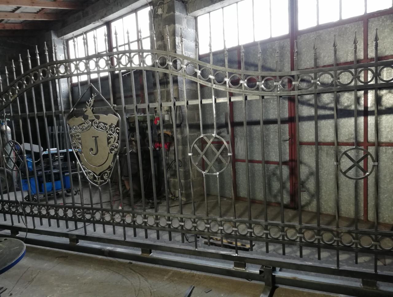 Surowa, metalowa konstrukcja bramy z dekoracyjnymi elementami, w tym herbem z literą 'J', w warsztacie z widocznym sprzętem spawalniczym i oknami w tle.