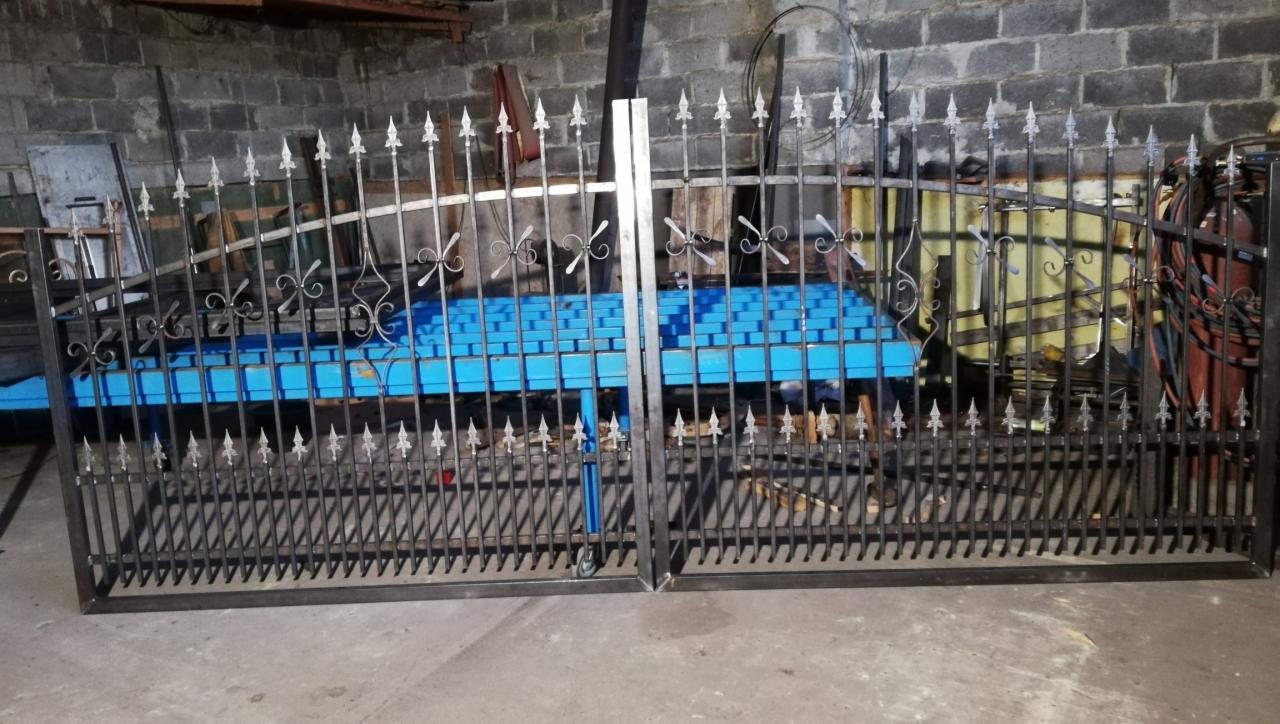 Surowa, spawana brama dwuskrzydłowa z kutymi zdobieniami w warsztacie, widoczne niebieskie profile stalowe w tle i ściana z szarej cegły.