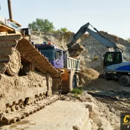 Ciężki sprzęt budowlany, w tym spychacz D5M LGP i koparka kołowa, pracują przy wykopie ziemnym z widoczną skarpą i zaparkowaną ciężarówką.