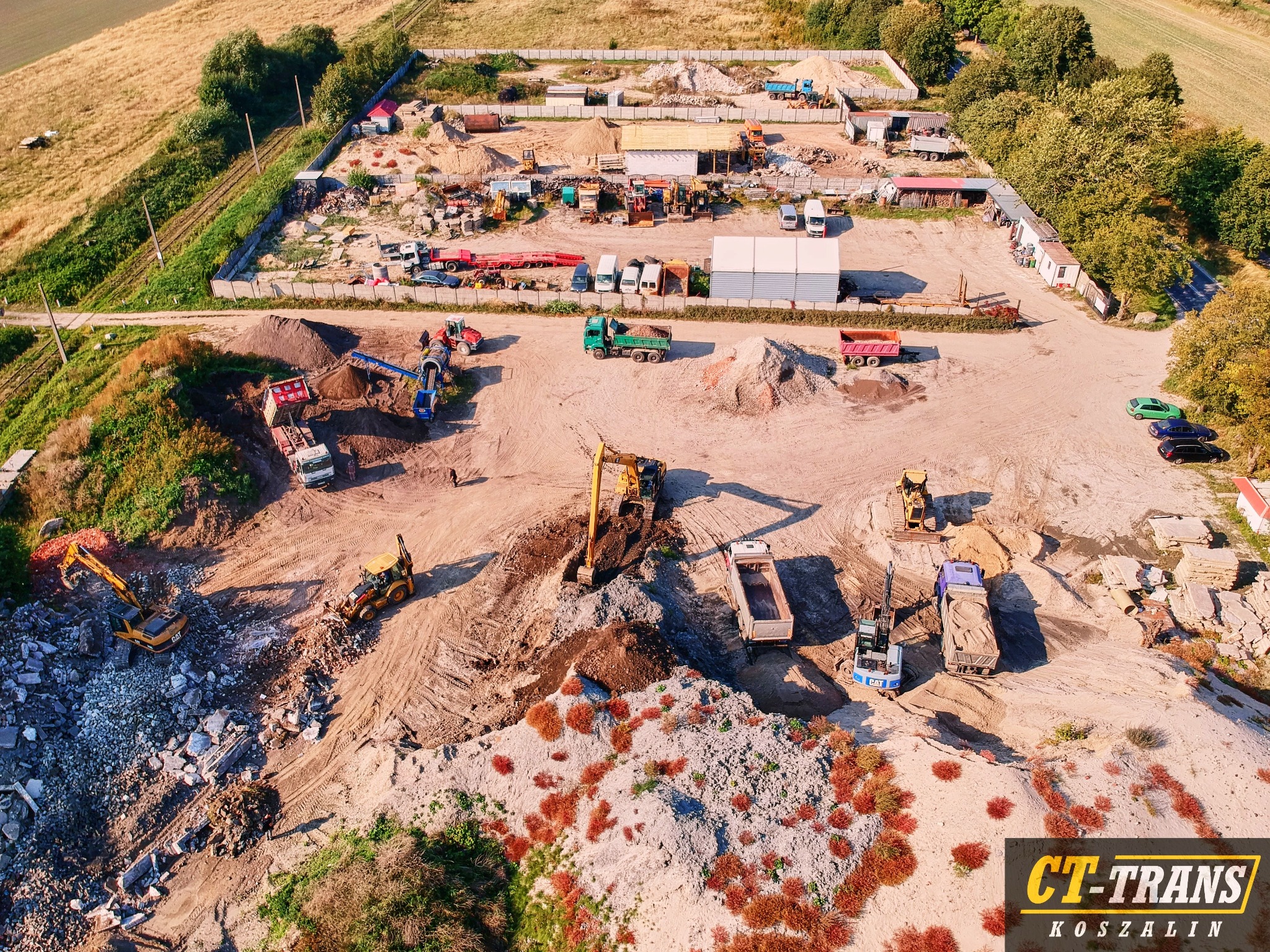 Szeroki kadr z lotu ptaka ukazuje plac budowy z licznymi maszynami, ciężarówkami i stertami ziemi, otoczony ogrodzeniem z prefabrykatów betonowych; logo firmy CT-Trans Koszalin w prawym dolnym rogu.