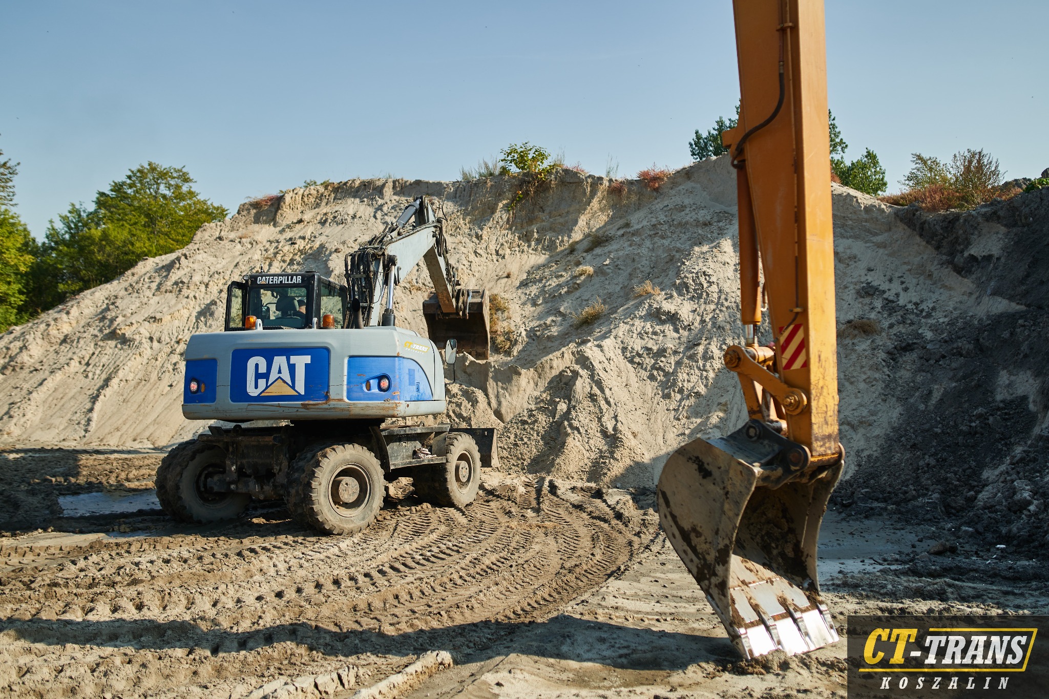 Kołowa koparka CAT z otwartą kabiną, pracująca przy kopcu piasku, widoczne ślady opon na piaszczystym podłożu, ramię koparki z łyżką w pierwszym planie.