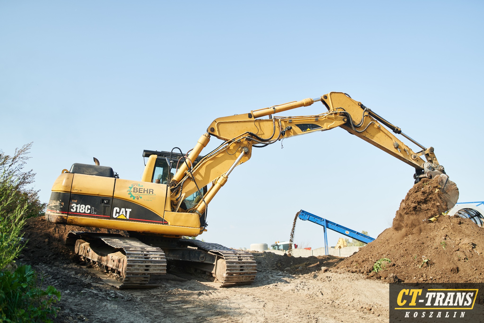 Żółta koparka CAT 318CL w trakcie prac ziemnych, wysypująca urobek na pryzmę ziemi na tle błękitnego nieba. Widoczny napis BEHR na maszynie i logo CT-TRANS w prawym dolnym rogu.