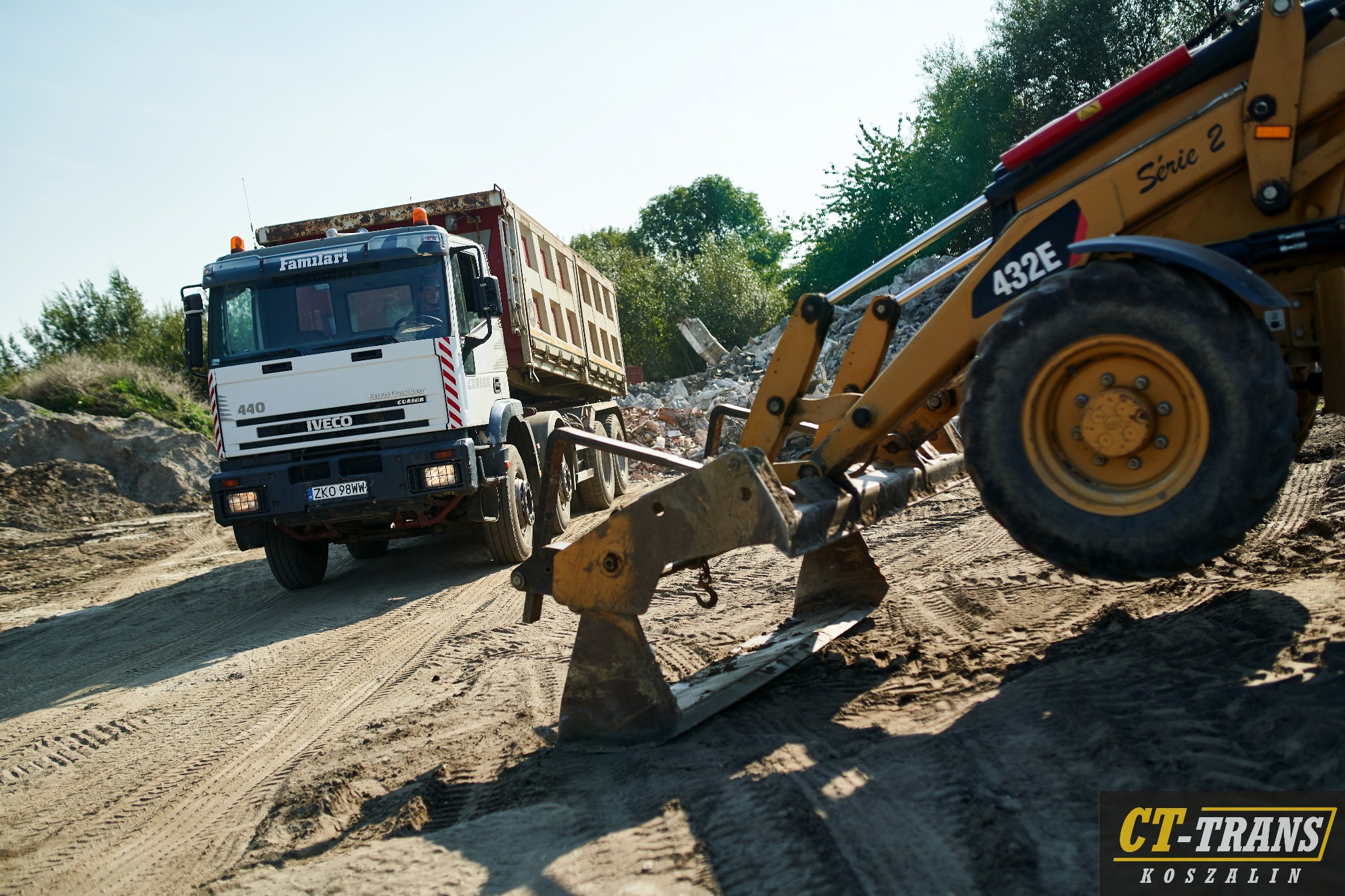 Ciężarówka Iveco z naczepą wywrotką na piaszczystej drodze, obok żółta koparko-ładowarka Case 432E Serie 2 z opuszczonym pługiem, ślady opon na piasku.