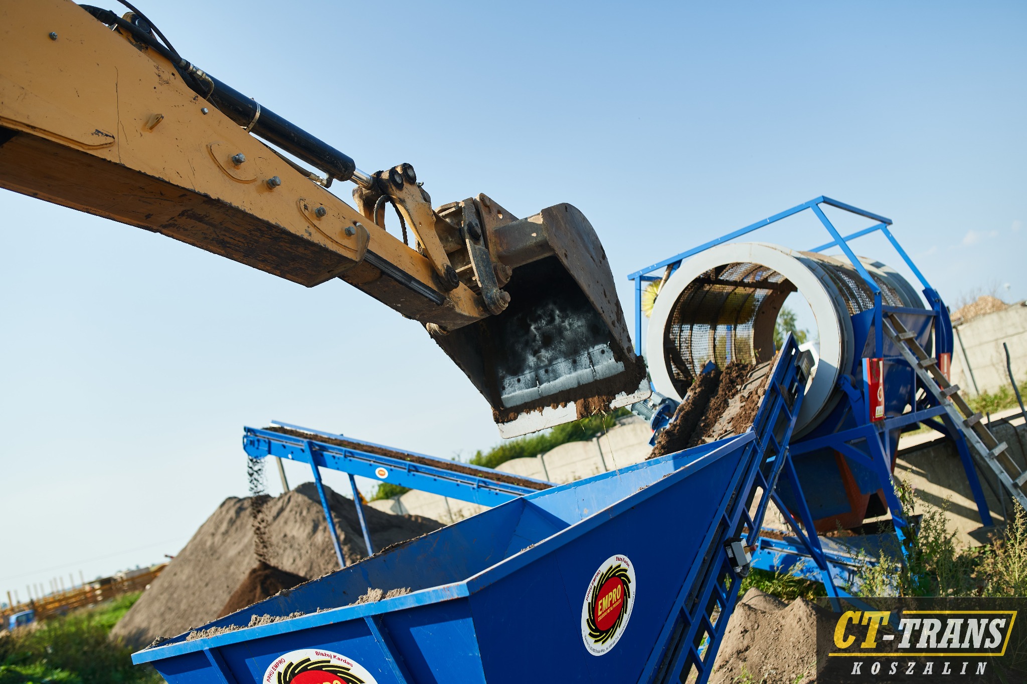 Żółta koparka z łyżką wysypuje ziemię do niebieskiego kontenera z logo firmy na tle przesiewacza bębnowego i błękitnego nieba.