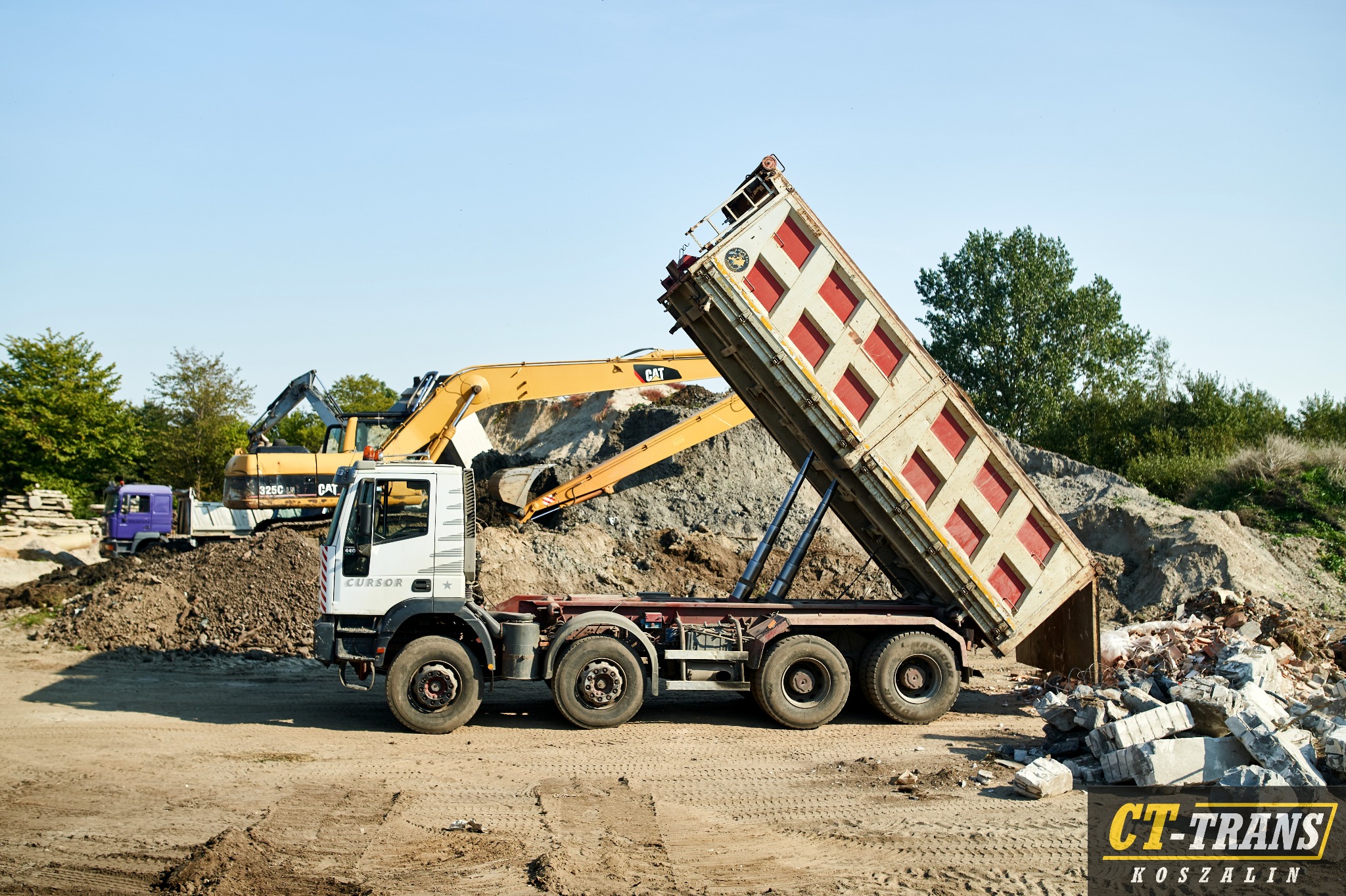 Biała ciężarówka z podniesioną skrzynią ładunkową, wysypująca gruz na ziemię. W tle żółta koparka CAT i inna ciężarówka. Widoczne ślady opon na piaszczystym podłożu. Na dole widoczne logo firmy.