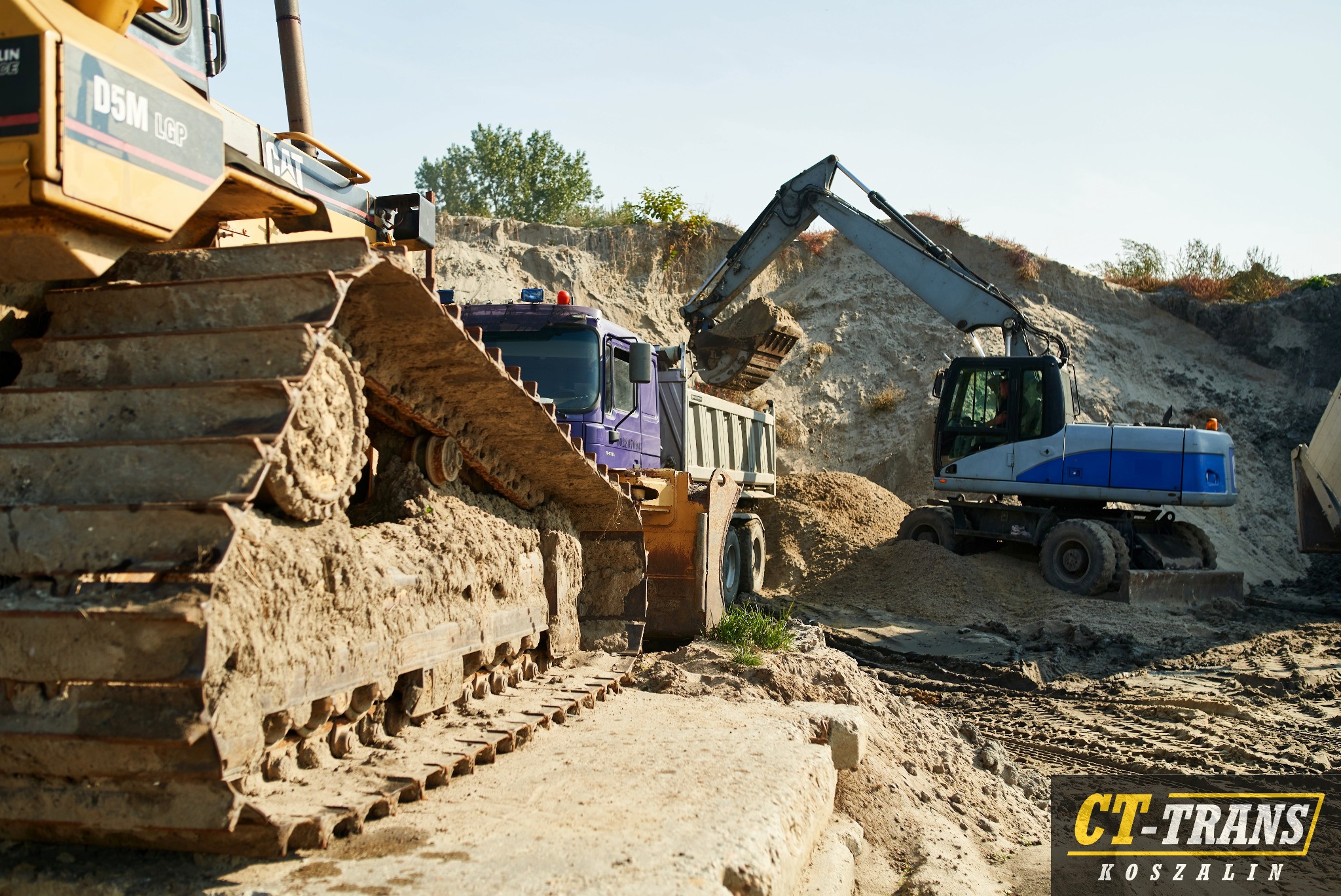 Ciężki sprzęt budowlany, w tym spychacz D5M LGP i koparka kołowa, pracują przy wykopie ziemnym z widoczną skarpą i zaparkowaną ciężarówką.