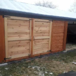 Drewniana konstrukcja z przesuwnymi drzwiami i żaluzjową ścianą boczną, z czarną rynną i śniegiem na dachu, na tle trawnika z resztkami śniegu.