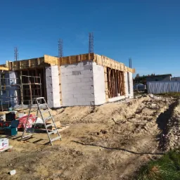 Budowa domu z bloczków, widoczne zbrojenia, szalunki, drabina, narzędzia i materiały budowlane w słoneczny dzień.