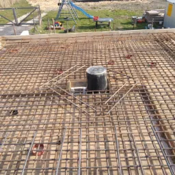 Zbrojenie płyty fundamentowej domu z widoczną instalacją kanalizacyjną i deskowaniem, w tle plac budowy z placem zabaw.