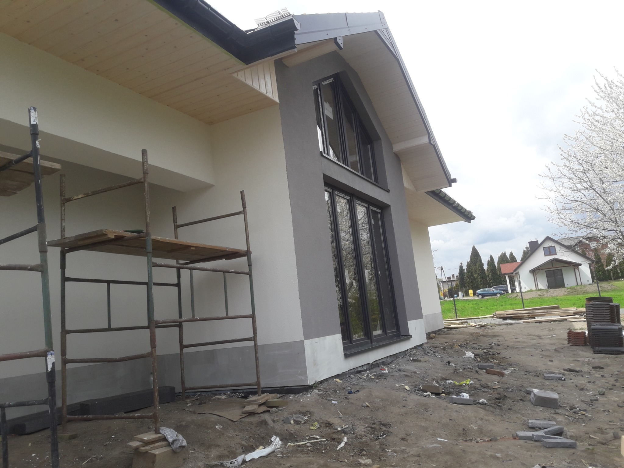 Nowoczesny dom jednorodzinny w trakcie budowy, z widocznym rusztowaniem, elewacją w odcieniach szarości i bieli, dużymi oknami i surowym otoczeniem.