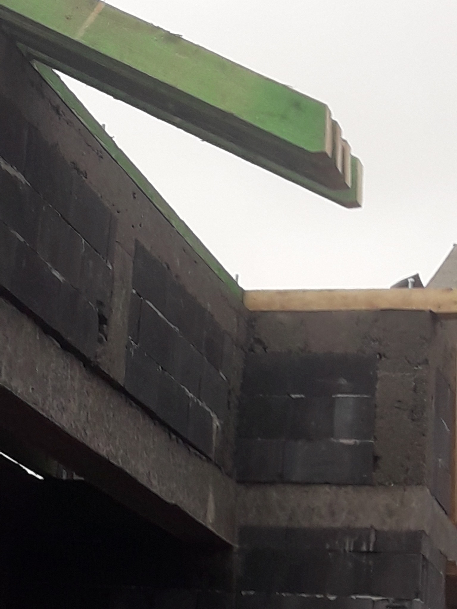 Wznoszenie ściany z ciemnoszarych bloczków betonowych, widoczny fragment zielonej belki stropowej i szare niebo w tle.
