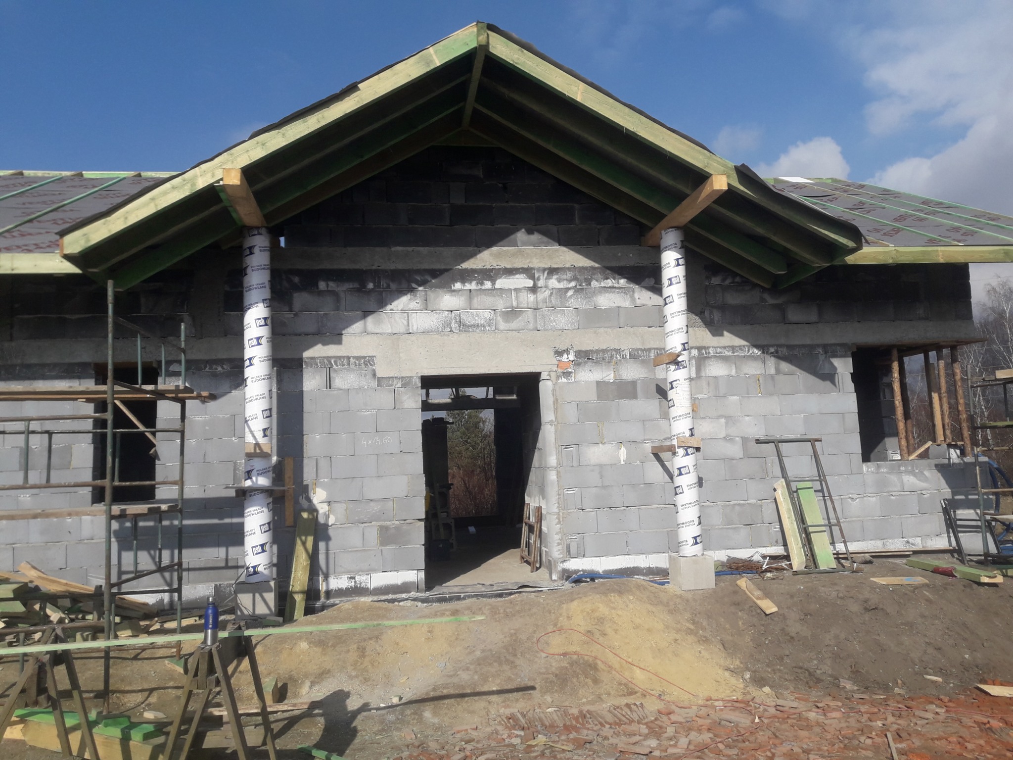 Surowy stan domu murowanego z bloczków betonowych, widoczne rusztowania, drewniana konstrukcja dachu z papą, słupy owinięte folią ochronną, teren budowy z gruzem i piaskiem.