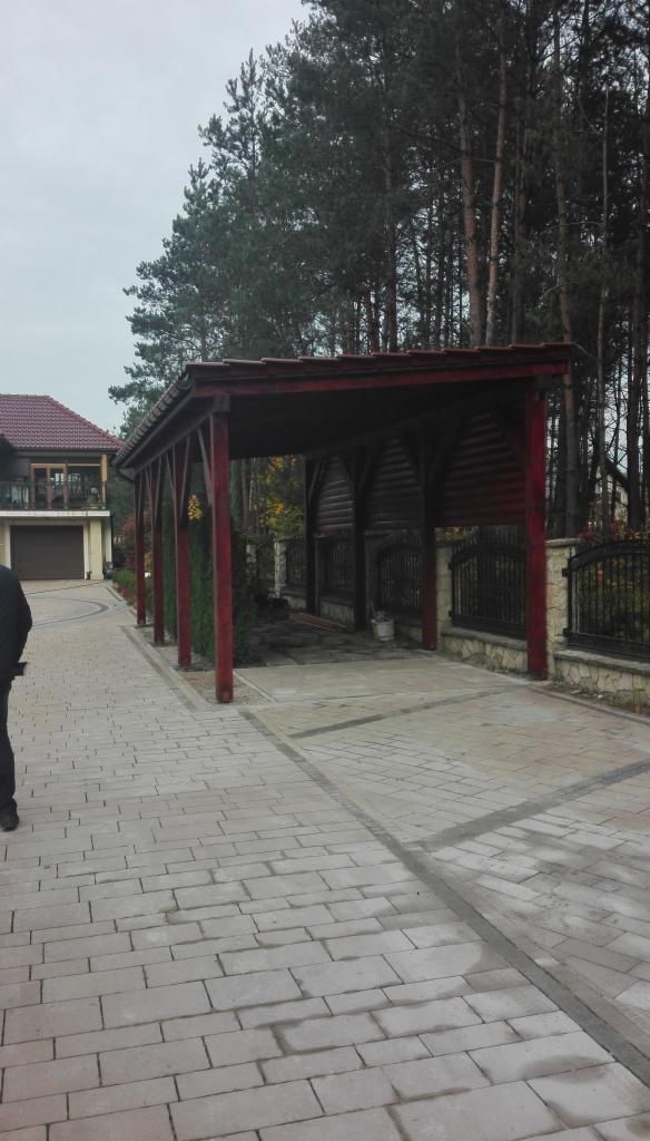 Drewniana wiata garażowa z czerwonymi słupami na posesji z kostki brukowej, widok na dom i las w tle.