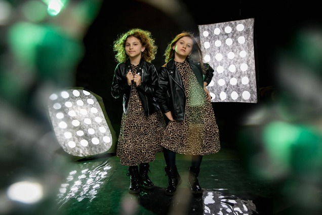 Dwie dziewczynki w identycznych sukienkach w panterkę i czarnych skórzanych kurtkach pozują na tle studyjnych lamp i czarnego tła.