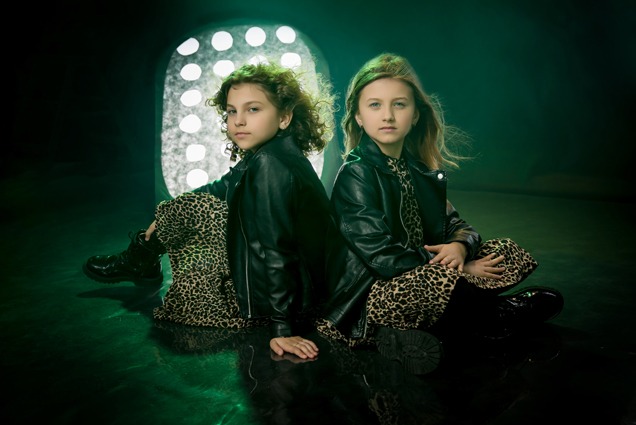 Dwie dziewczynki w czarnych skórzanych kurtkach i panterkowych strojach siedzą plecami do siebie na błyszczącej, ciemnej powierzchni, oświetlone zielonym światłem i reflektorem.