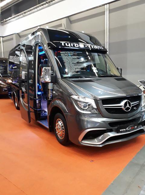 Szary, tuningowany bus marki Mercedes z otwartymi drzwiami i podświetleniem wnętrza, zaparkowany na pomarańczowej podłodze prawdopodobnie na targach transportowych w Wołowie. Na pojeździe widoczny...