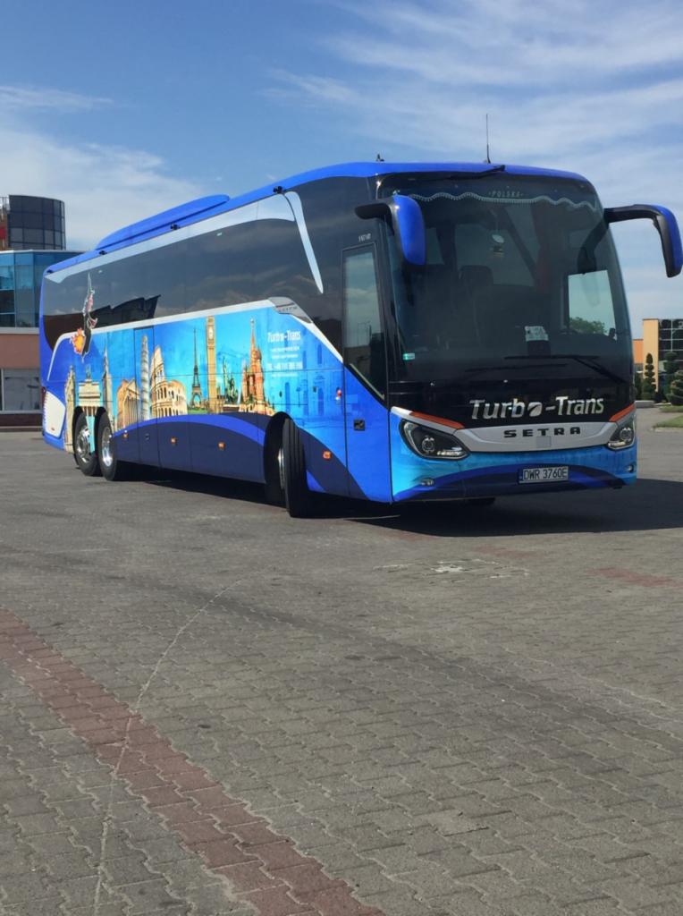 Niebieski autokar Setra Turbo-Trans z panoramą europejskich miast na boku, zaparkowany na placu z kostki brukowej.