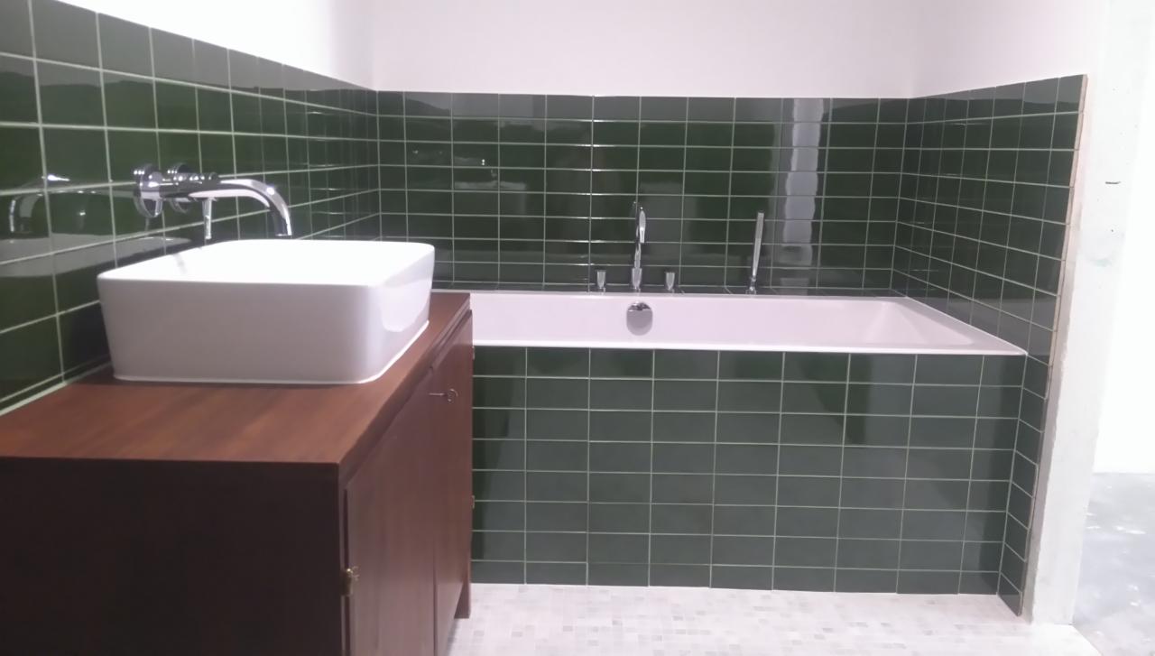 Nowoczesna łazienka z wanną obudowaną ciemnozielonymi, błyszczącymi płytkami, białą, prostokątną umywalką nablatową na szafce z ciemnego drewna oraz mozaikową podłogą w odcieniach szarości.