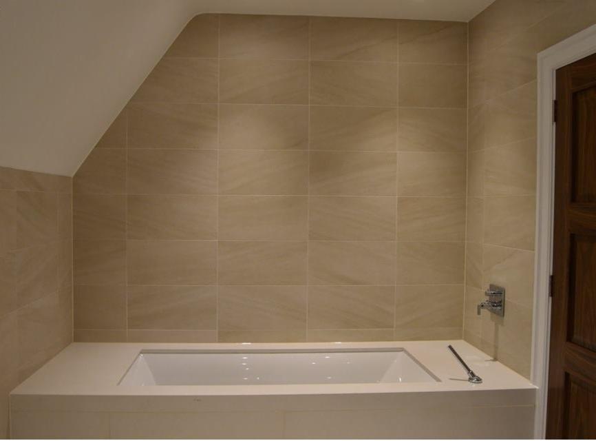 Beżowe płytki ceramiczne na ścianie w łazience z narożną wanną obudowaną płytkami, widoczna bateria wannowa i drewniane drzwi